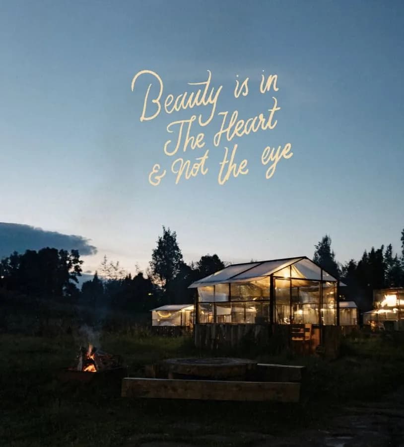 Casa de vidrio iluminada al atardecer con un letrero de neón que dice “Beauty is in the heart & not the eye”